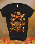 Black t-shirt with a graphic of a person with a bun, glasses, and leaves with 'Grateful' text on a blurred autumn background.